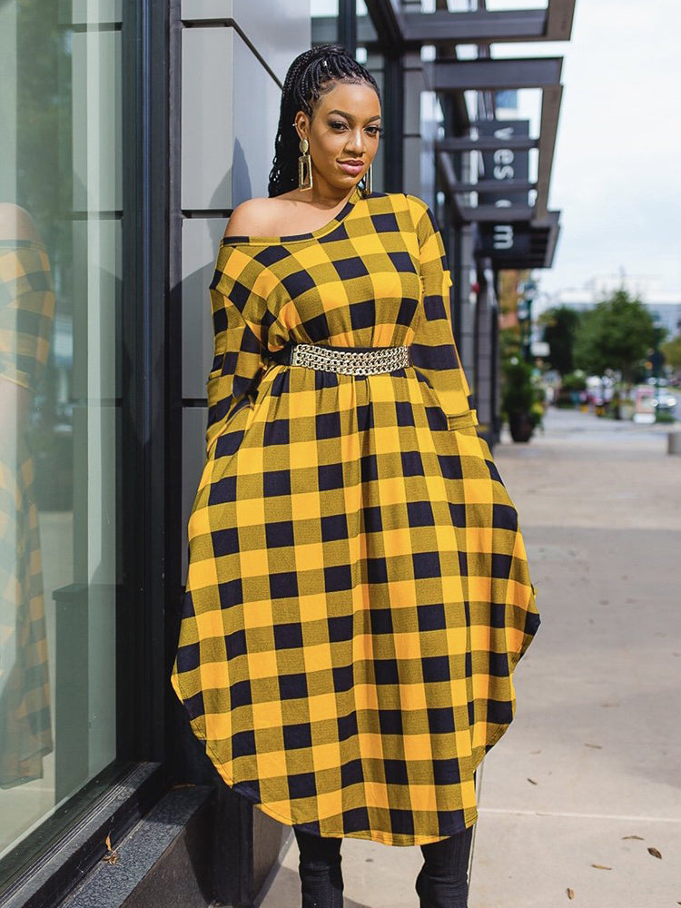 A woman stands on a sidewalk wearing a yellow and black plaid off-the-shoulder midi dress cinched with a gold belt.