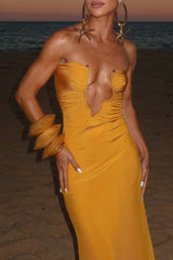 Close-up of a woman's upper body in a yellow strapless gown with a deep V-neckline and ruched detailing, accessorized with statement jewelry, on a beach.