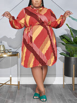 Plus-size woman wearing a warm-toned, long-sleeved, pleated A-line dress with a geometric print and tie-waist.
