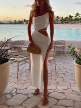 Woman in a white ruched one-shoulder dress with cut-out details and a high slit, posing on a patio by the ocean.
