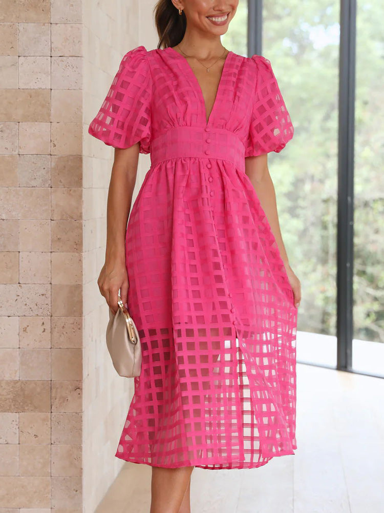 A woman smiling and holding the hem of her pink midi dress, which features a textured grid overlay and a V-neckline.