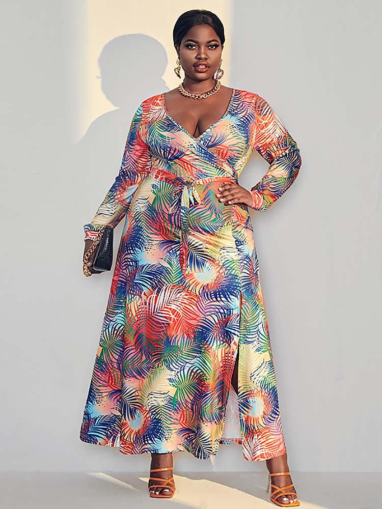 Woman wearing a colorful tropical print maxi dress with long sleeves and a V-neck wrap design, standing against a light gray wall.