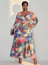 Woman wearing a colorful tropical print maxi dress with long sleeves and a V-neck wrap design, standing against a light gray wall.