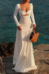Woman wearing a white long-sleeved maxi dress with a cut-out and ruched detailing, standing by the sea.