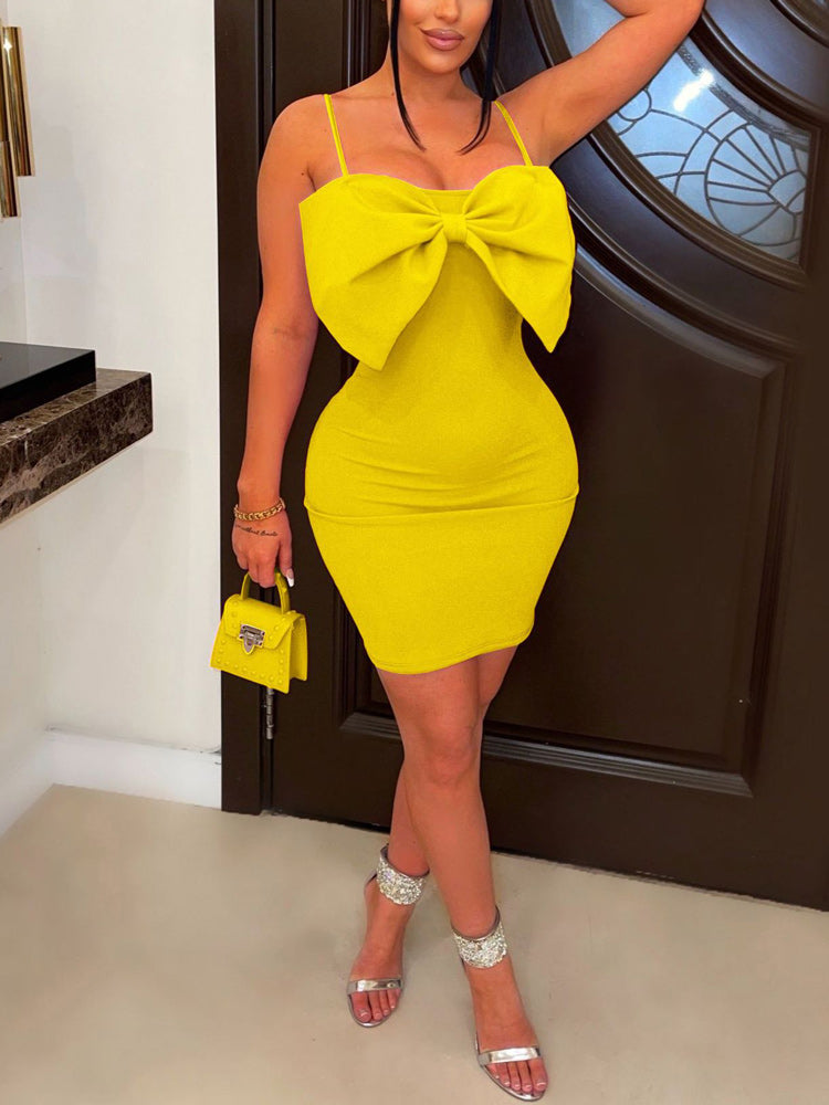 Woman wearing a bright yellow mini dress with a large bow detail on the bust and spaghetti straps, paired with silver heels and a small yellow handbag.