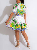 Rear view of a woman in a white short-sleeve dress with a tropical fruit and leaf print, showing the pleated skirt and lace-up heels.
