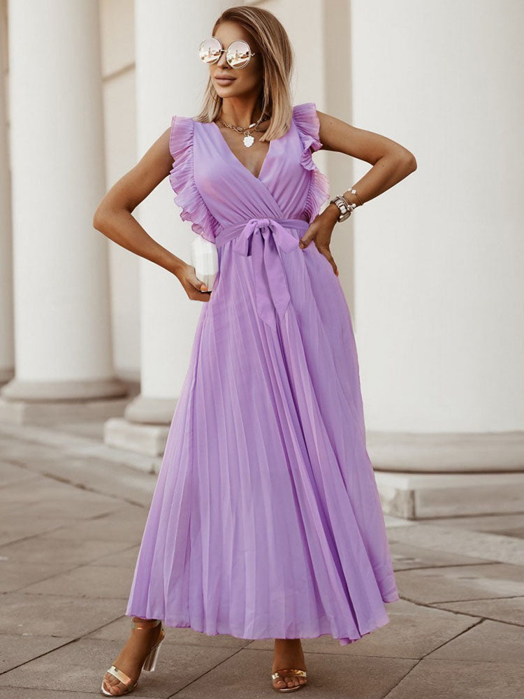 woman posing in a long purple pleated dress with ruffled sleeves and a tie belt, wearing sunglasses and holding a clutch