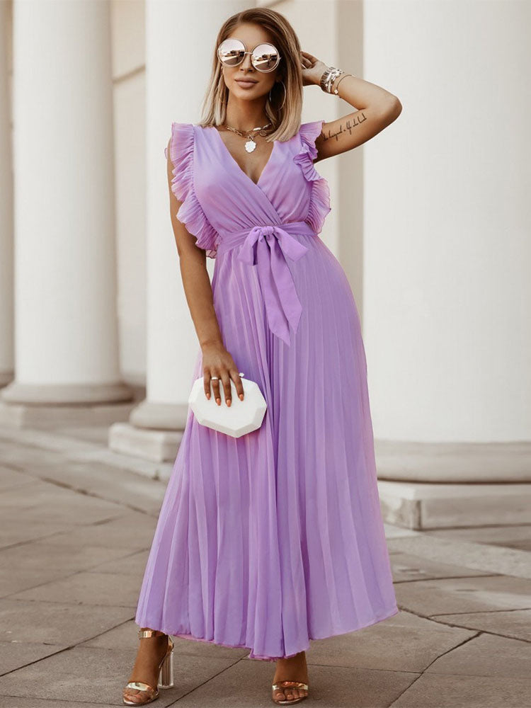 woman posing in a long purple pleated dress with ruffled sleeves and a tie belt, wearing sunglasses and holding a clutch