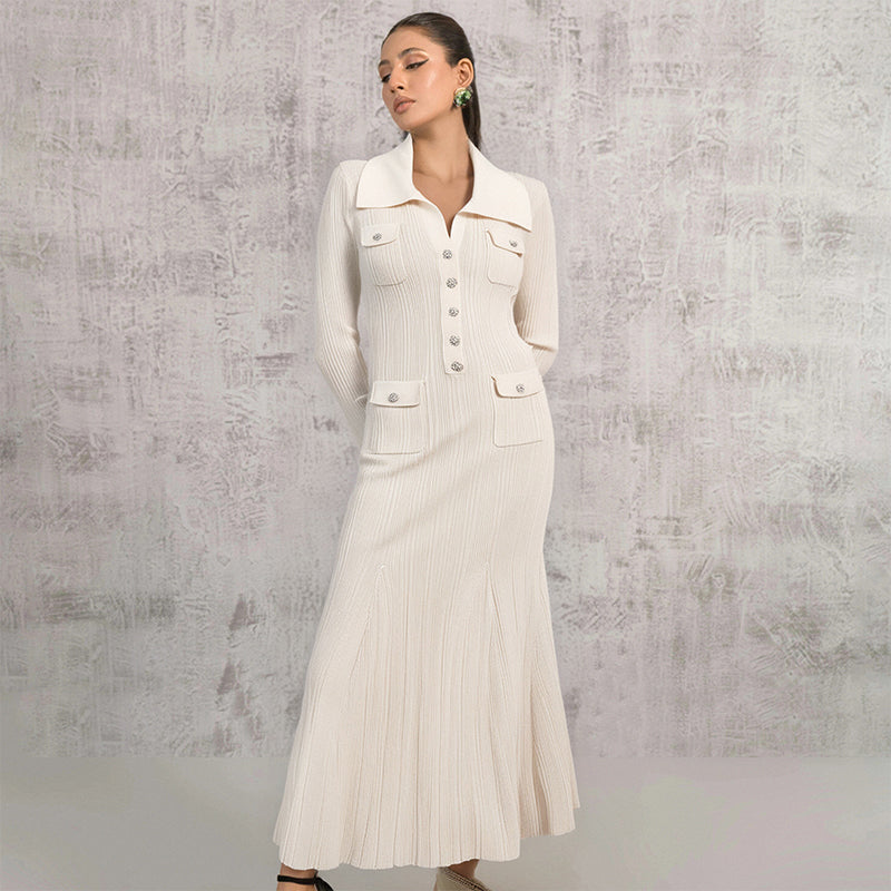 Woman wearing a cream-colored long-sleeved knit dress with a collared neckline, button-up front, dual chest pockets, and a flared hem, standing against a textured wall.