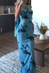 A woman poses in a room wearing a blue floral one-shoulder maxi dress.