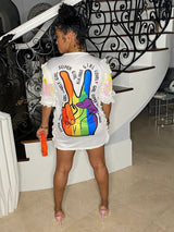 Back view of a woman wearing a white sequined t-shirt dress with a rainbow peace sign graphic and text.