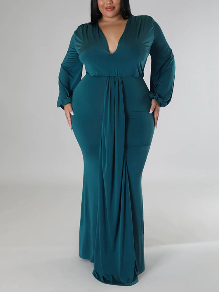 Woman wearing a teal, floor-length, long-sleeved gown with a V-neckline and draped skirt, standing with hands on hips