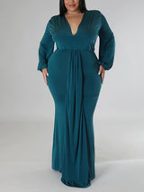 Woman wearing a teal, floor-length, long-sleeved gown with a V-neckline and draped skirt, standing with hands on hips
