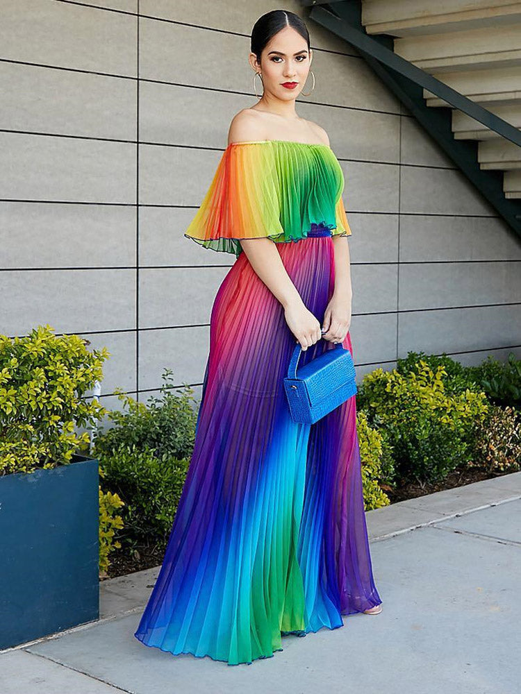 Woman wearing a rainbow gradient off-the-shoulder pleated maxi dress and holding a blue clutch, standing outdoors with a modern building in the background.