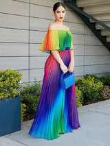 Woman wearing a rainbow gradient off-the-shoulder pleated maxi dress and holding a blue clutch, standing outdoors with a modern building in the background.
