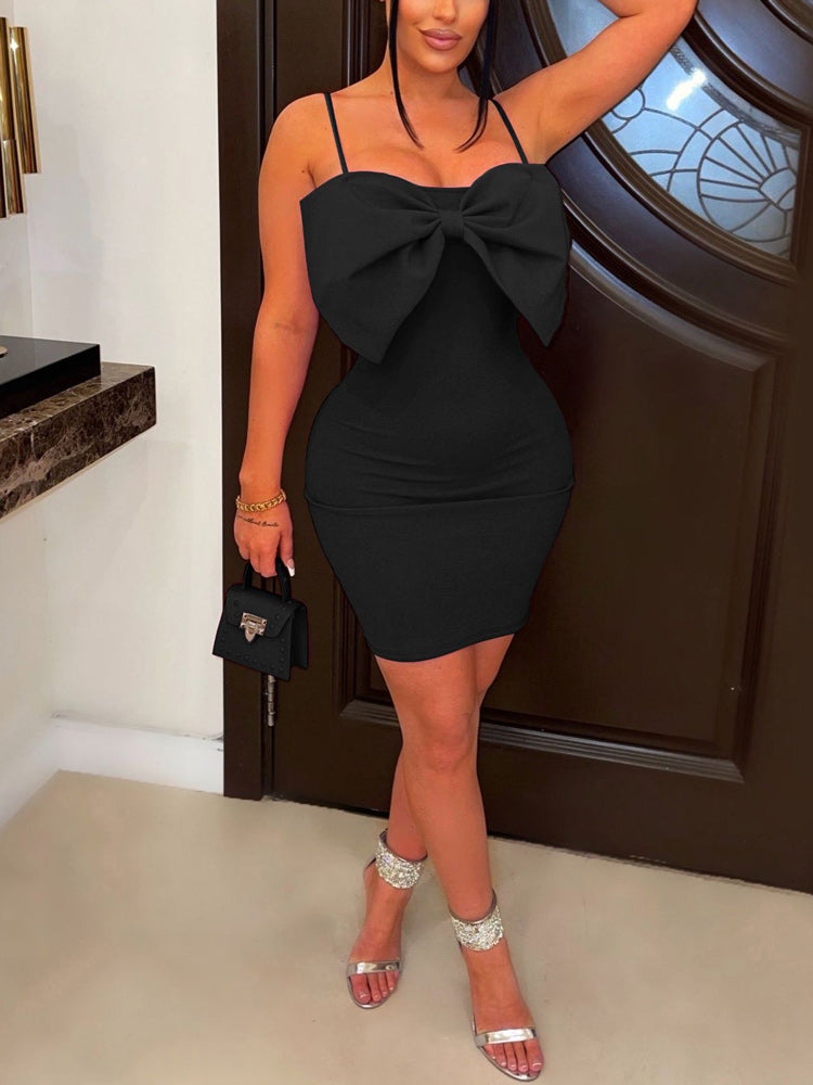 Woman wearing a black mini dress with a large bow detail on the bust and spaghetti straps, paired with silver heels and a small black handbag.