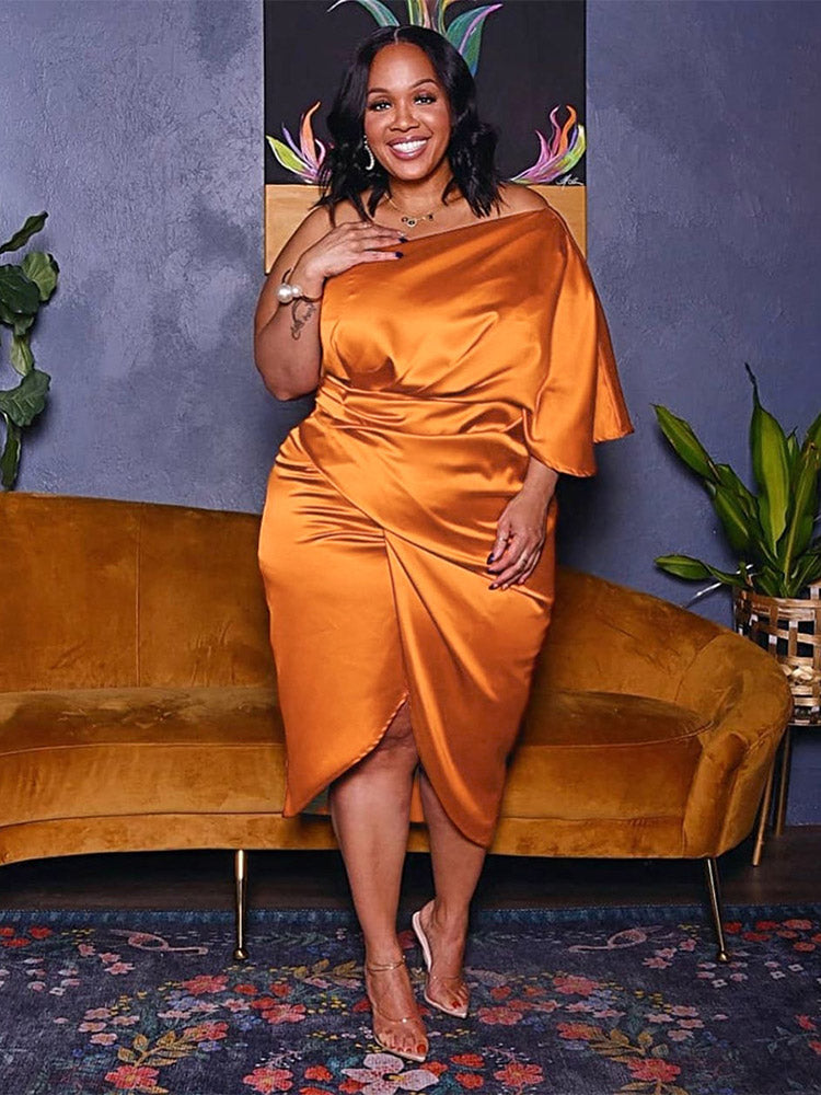 Woman wearing an orange satin one-shoulder dress with a draped bodice and tulip hem skirt, standing in front of a mustard yellow couch.