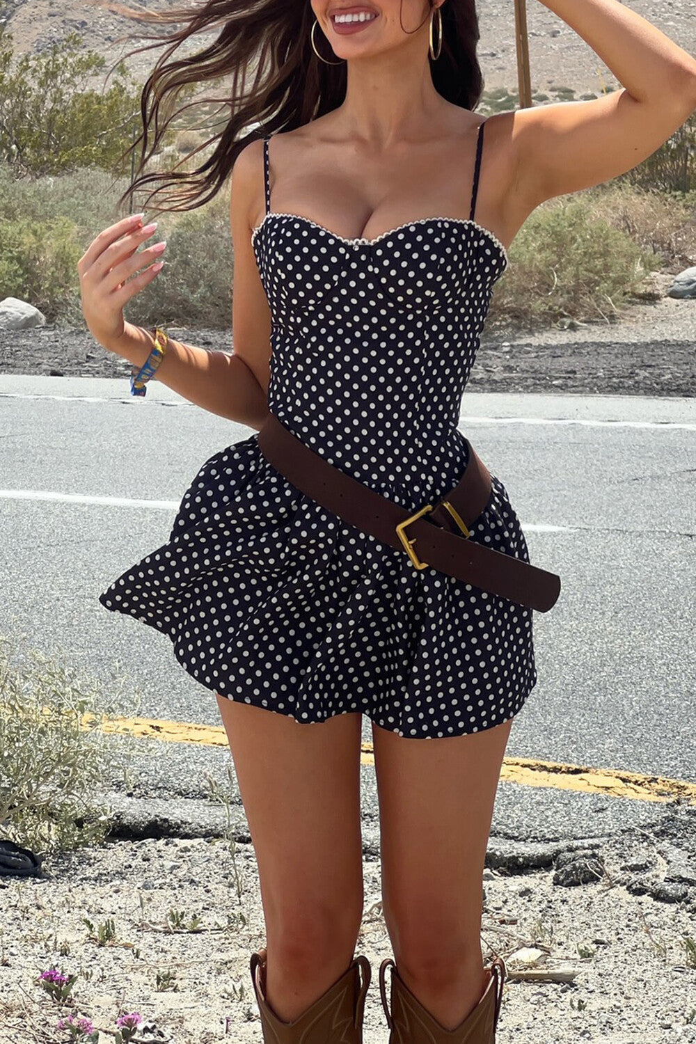 Woman with windblown hair wearing a polka dot mini dress and brown belt.