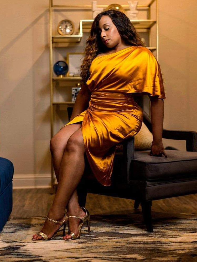 Woman sitting on a chair wearing a gold satin one-shoulder dress with a draped bodice and tulip hem skirt.