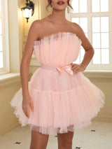 Woman wearing a short, strapless, tiered tulle dress in a soft pink color, with ruffled detailing and a satin bow at the waist.