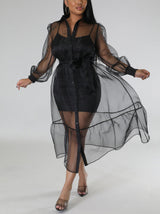 Woman wearing a sheer black organza dress over a black slip dress, belted at the waist with puff sleeves and a tiered hem.