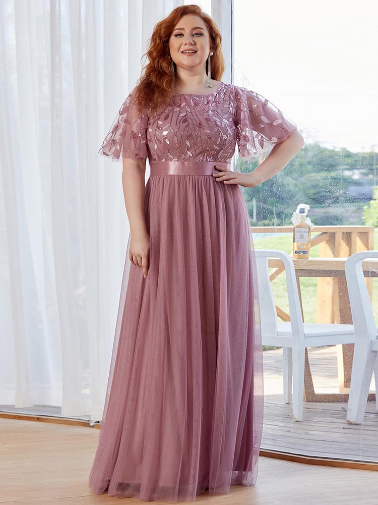 Woman with auburn hair wearing a dusty rose A-line maxi dress with embroidered flutter sleeves and a satin waistband, posing indoors.