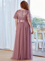Back view of a woman wearing a dusty rose A-line maxi dress with embroidered flutter sleeves and a satin waistband.