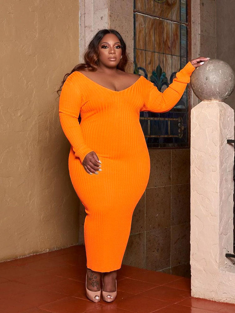 Woman in an orange ribbed knit midi dress with long sleeves and an off-the-shoulder V-neckline, posing with hands on hips.