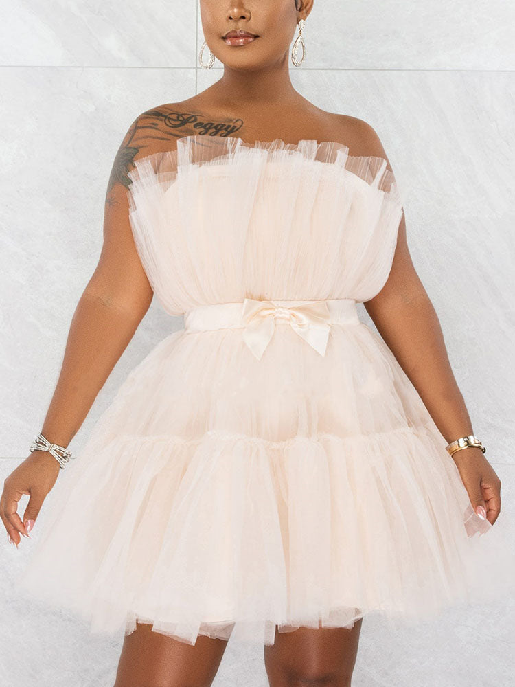 Woman wearing a short, strapless, tiered tulle dress in a light peachy-pink color, with a satin bow at the waist.