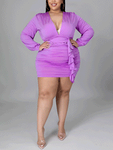 Plus-size woman wearing a purple ruched mini dress with a v-neck and long sleeves, standing with hands on hips.