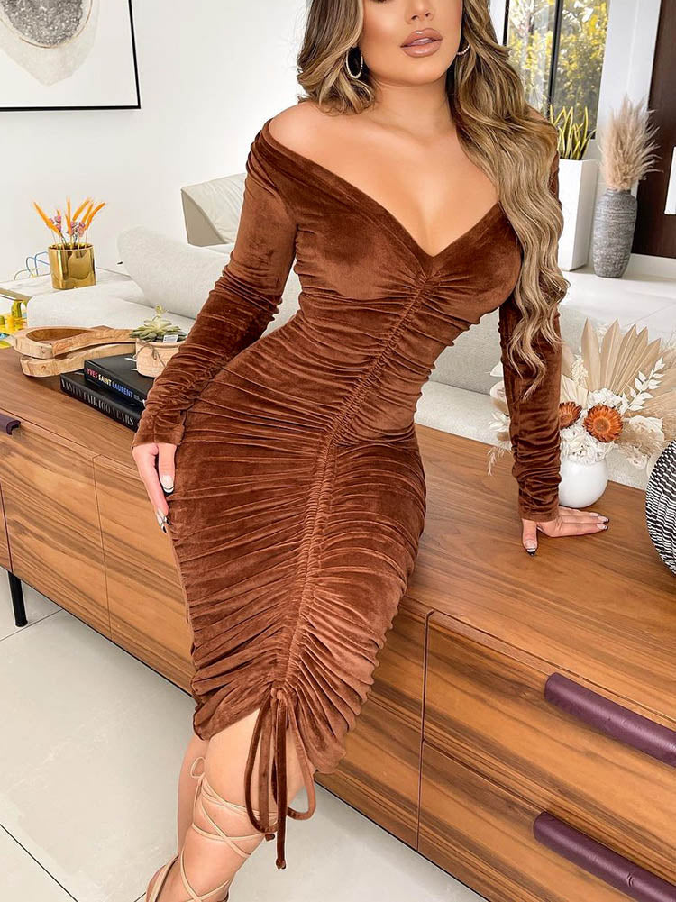 Woman wearing a brown off-the-shoulder ruched velvet dress with long sleeves and a drawstring hem, posing leaning on a wooden cabinet.