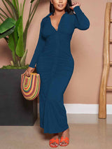 Woman wearing a teal ruched maxi dress with a collared v-neck and long sleeves, holding a colorful woven bag.