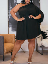 Plus-size woman in a black one-shoulder dress with an asymmetrical sleeve, hands on hips, in a living room setting.