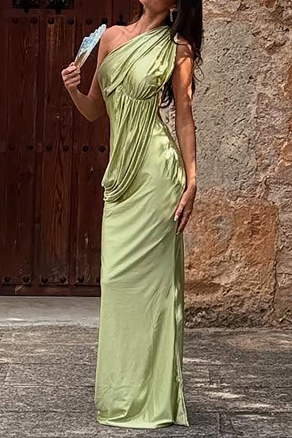 Woman in a lime green satin one-shoulder maxi dress with draped detailing, standing in front of a wooden door.