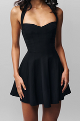 Torso of a woman in a black halter neck mini dress with sweetheart neckline and pleated skirt, against a grey background.