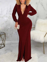 Woman wearing a long burgundy dress with a deep V-neckline, long sleeves, ruched detailing, and a high slit.