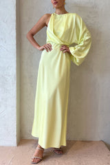 Woman wearing a pale yellow satin one-shoulder maxi dress with a draped bodice and one long asymmetrical sleeve, standing against a textured white wall.