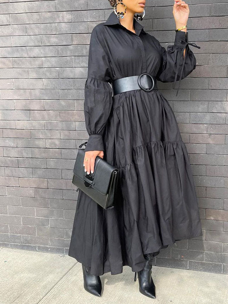 Woman wearing a black tiered maxi dress with balloon sleeves and a wide black belt, holding a black clutch bag.