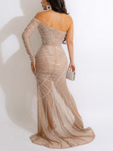 Back view of a woman in a nude, off-the-shoulder, floor-length gown with a high slit and silver embellishments, holding a silver clutch.