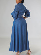 Back view of a woman in a blue denim maxi dress with puffed sleeves and a belted waist.