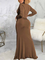 Back view of a woman wearing a long brown dress with a deep V-neckline, long sleeves, ruched detailing, and a high slit.
