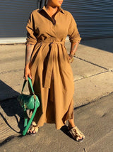 Woman wearing a long brown collared dress with a belted waist and a green handbag.