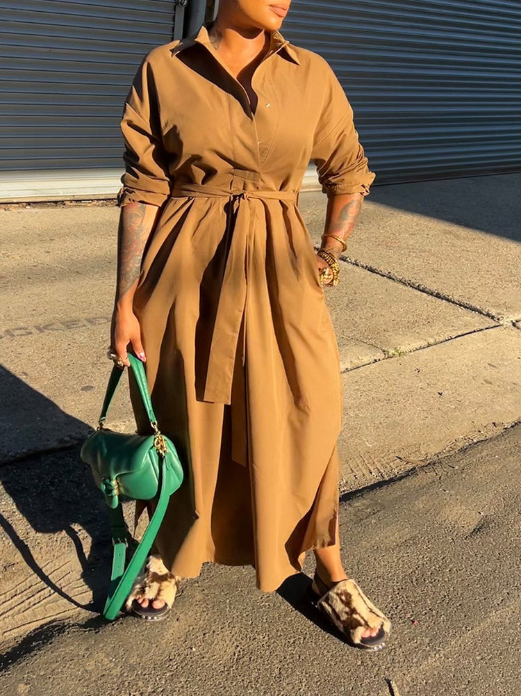 Woman wearing a long brown collared dress with a belted waist and a green handbag.