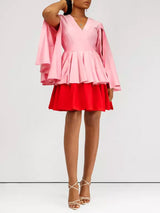 Woman in a pink and red color-block dress with cape sleeves and white strappy heels, posing against a white background.