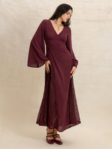 Woman in a burgundy maxi dress with flared sleeves and lace skirt panels, looking down elegantly.