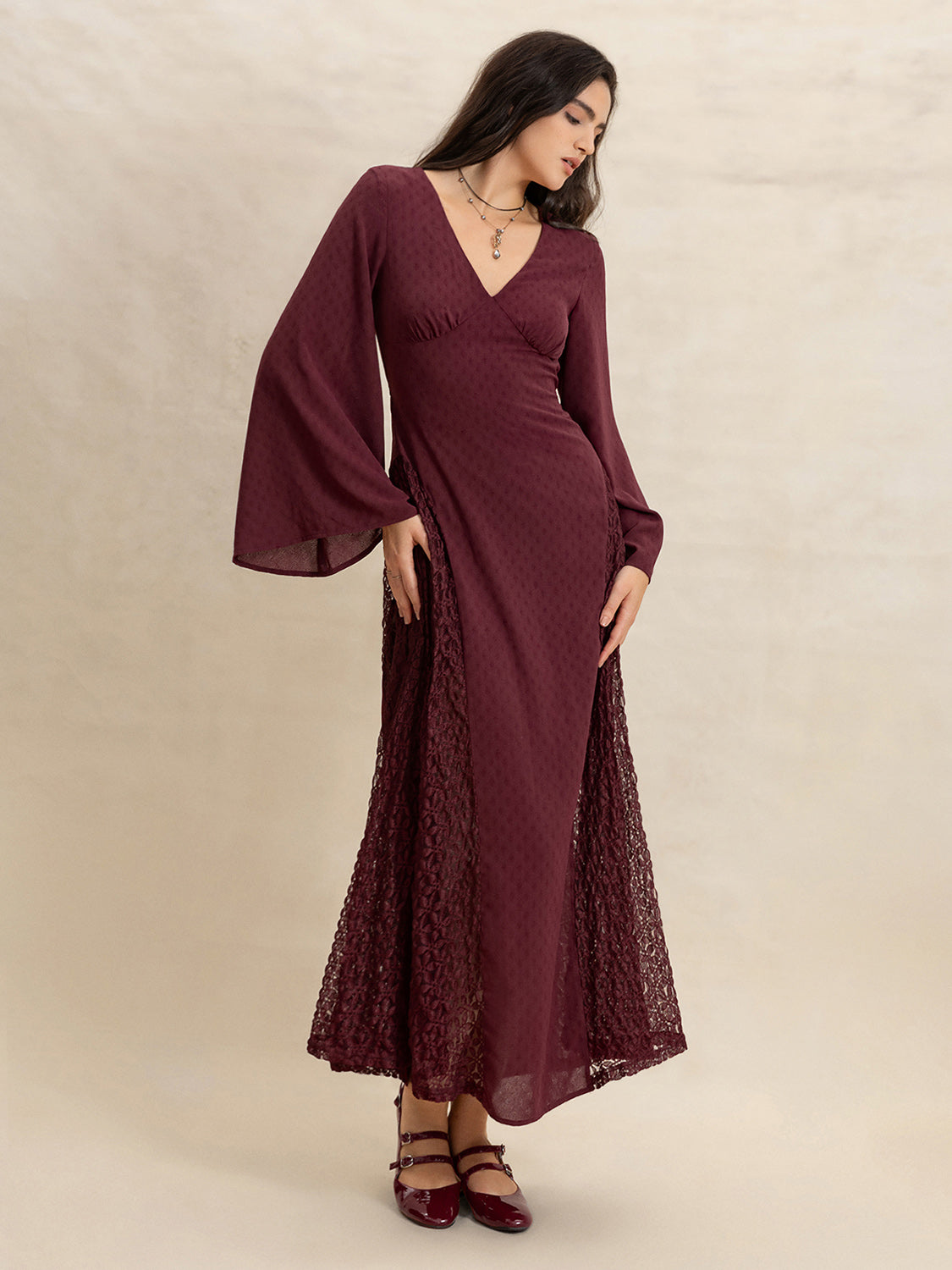Woman in a burgundy maxi dress with flared sleeves and lace skirt panels, looking down elegantly.