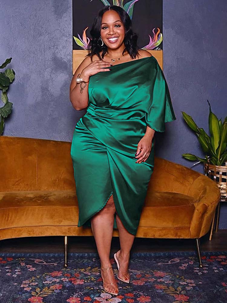 Woman in a green one-shoulder satin dress with draping and a tulip hem, smiling and touching her chest.