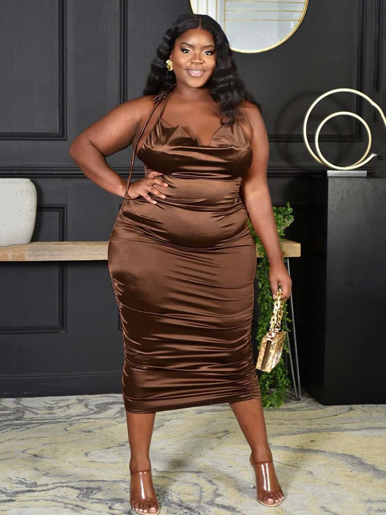 Woman wearing a brown satin ruched halter neck sheath dress and clear heeled sandals, holding a gold handbag.