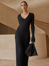 Woman wearing a black ribbed knit maxi dress with flared cuffs and a gold necklace, holding a black handbag.