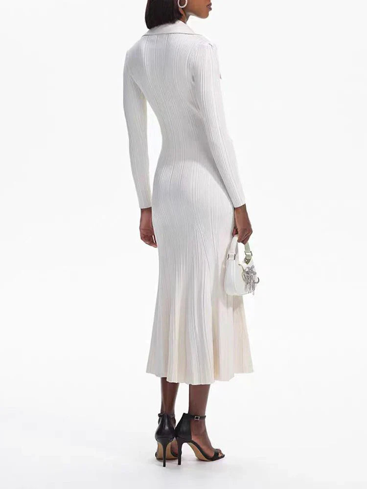 Back view of a woman wearing a cream-colored long-sleeved knit dress with a collared neckline, button-up front, dual chest pockets, and a flared hem, holding a white handbag and wearing black heels.